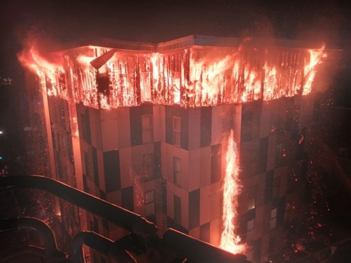 A large building engulfed in flames with fire streaming down from the roof and around the exterior walls at night, with visible sparks and embers highlighting the intensity of the blaze