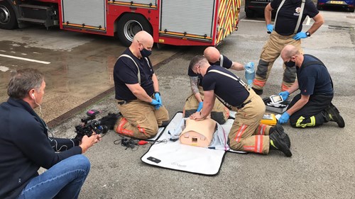Four firefighters training on CPR techniques using a lifelike dummy, while a man with a camera observes