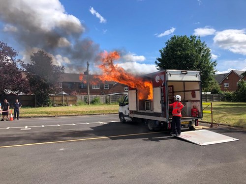 Flames coming out of kitchen safety unit demonstrating how a cooking fire can break out. Firefighter stood to the right in orange uniform and white helmet.