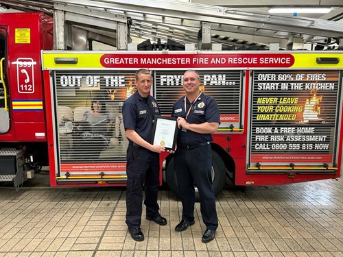 Two firefighters stand in front of a fire engine inside a fire station. One hands a certificate to the other