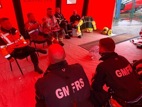 In a tent with red walls, the GMFRS crew sit on chairs planning their exercise