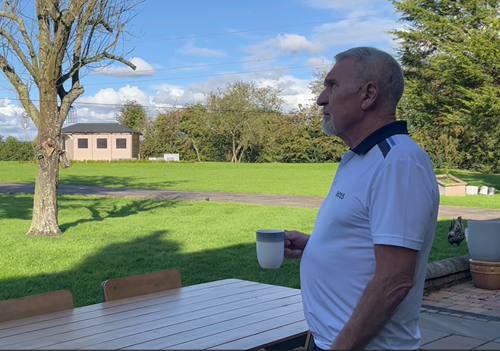 A person in a polo shirt holding a cup of tea in their garden.
