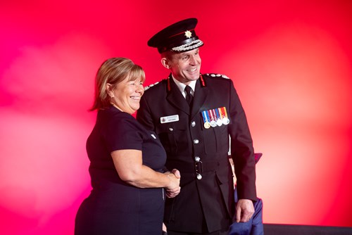 Chief Fire Officer, Dave Russel shaking hands with award recipient Seonaid Kendall during the presentation of her 30-year award clasp. The two are facing frame right and both are smiling, posing for a photograph.