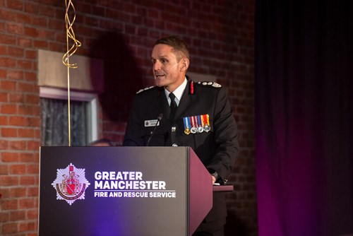 Chief Fire Officer, Dave Russel stood at a lectern with the GMFRS logo on the front. He is addressing a crowd in the room.