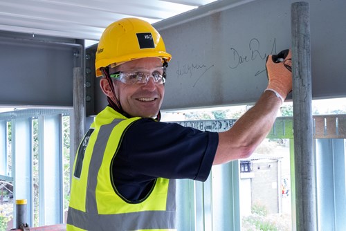 Man smiling, wearing high visibility jacket and hard helmet whilst signing a steel beam