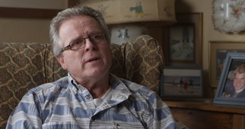 Nich Cunliffe sat on sofa talking to the camera with family photographs in the background