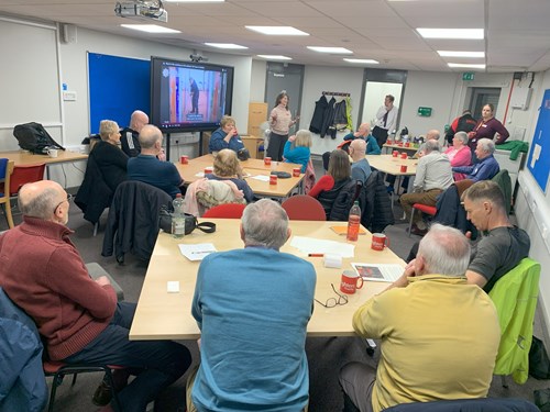 Living well group members sat around tables with refreshments listening to a speaker stood by a screen